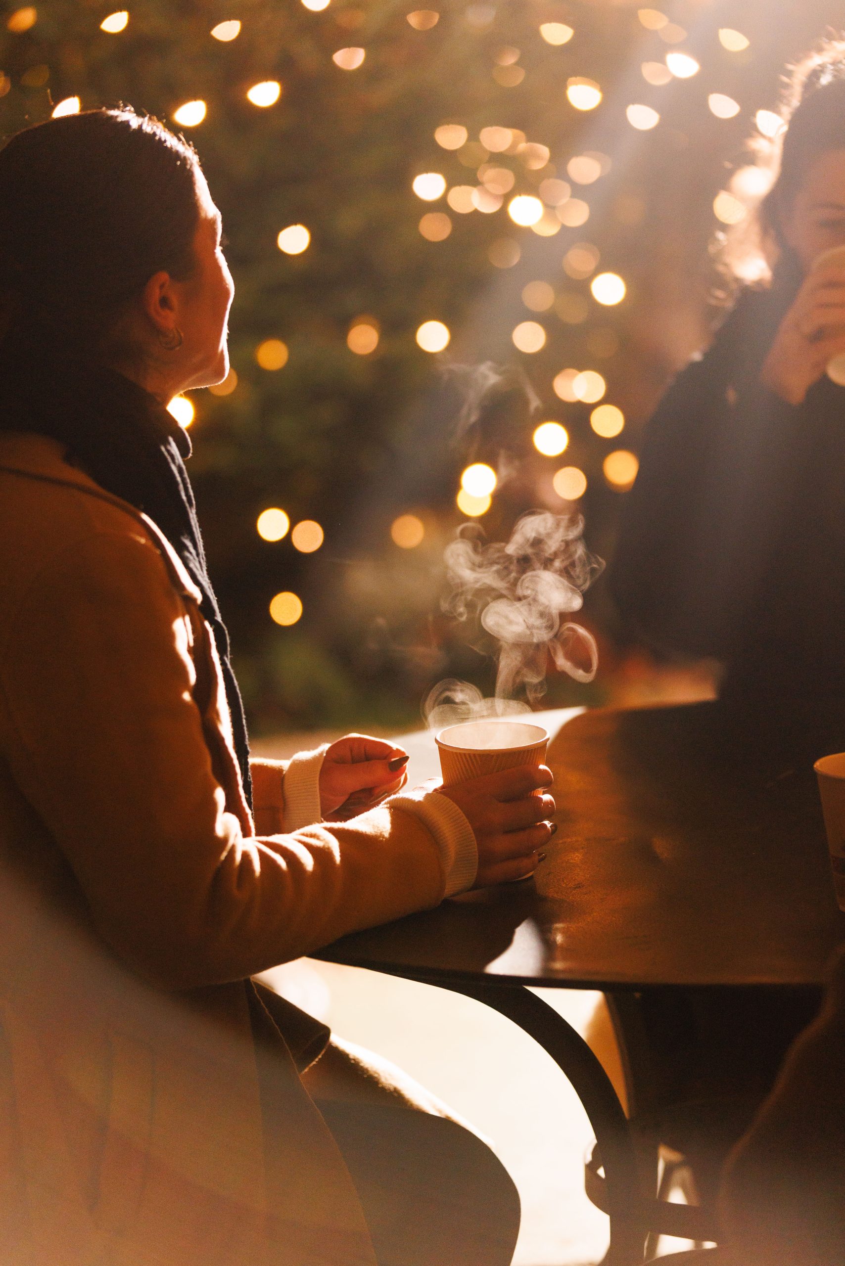 Eine Person sitzt an einem Tisch im Freien und hält eine dampfende Tasse in der Hand, während im Hintergrund warme Bokeh-Lichter leuchten. Eine weitere Person ist teilweise sichtbar und schafft eine gemütliche, festliche Atmosphäre.