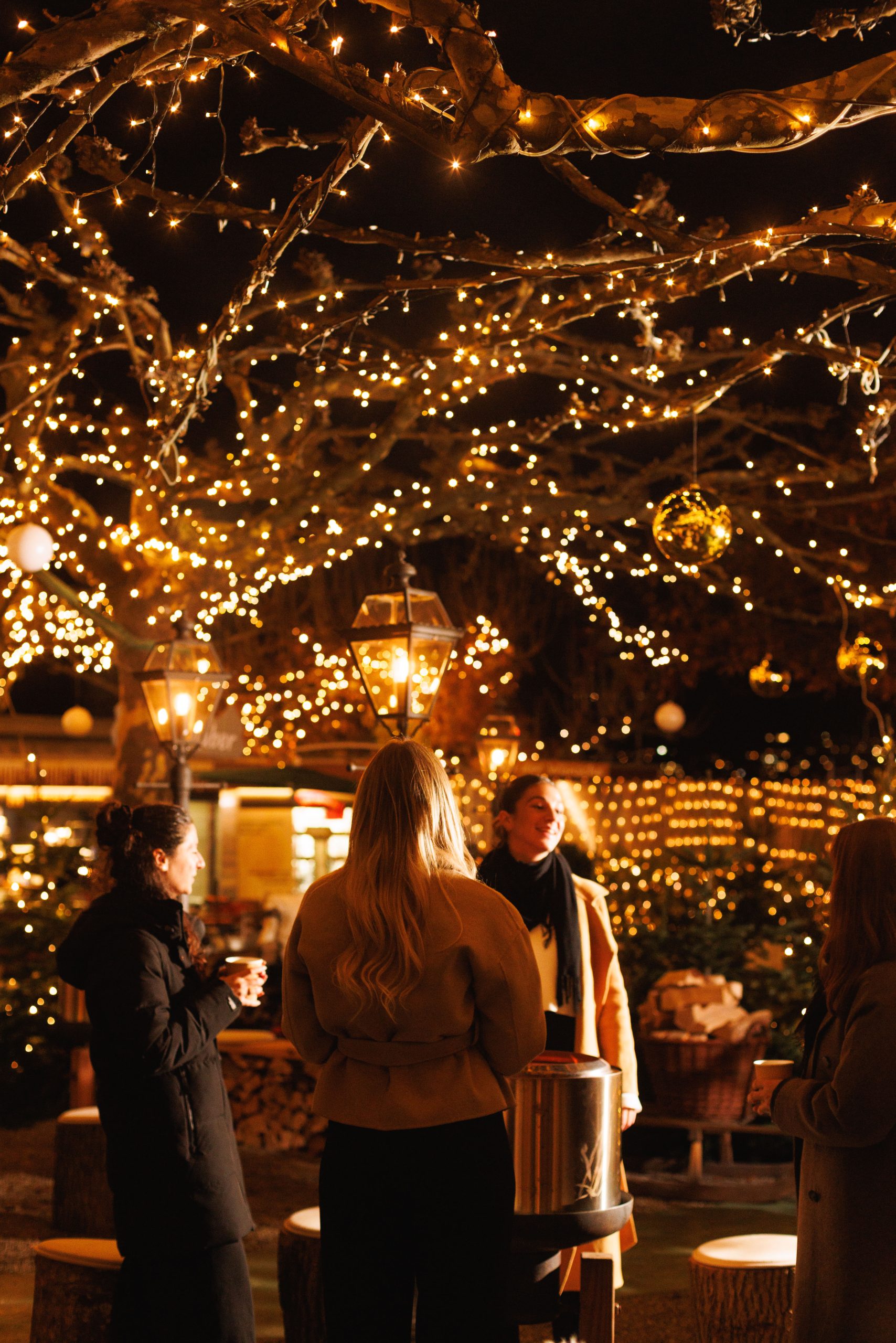 Vier Personen stehen nachts unter Bäumen, die mit warmen Lichterketten und hängenden Ornamenten geschmückt sind, und genießen eine festliche Zusammenkunft im Freien. Eine Person steht bei einem großen Topf, und alle scheinen sich zu unterhalten.