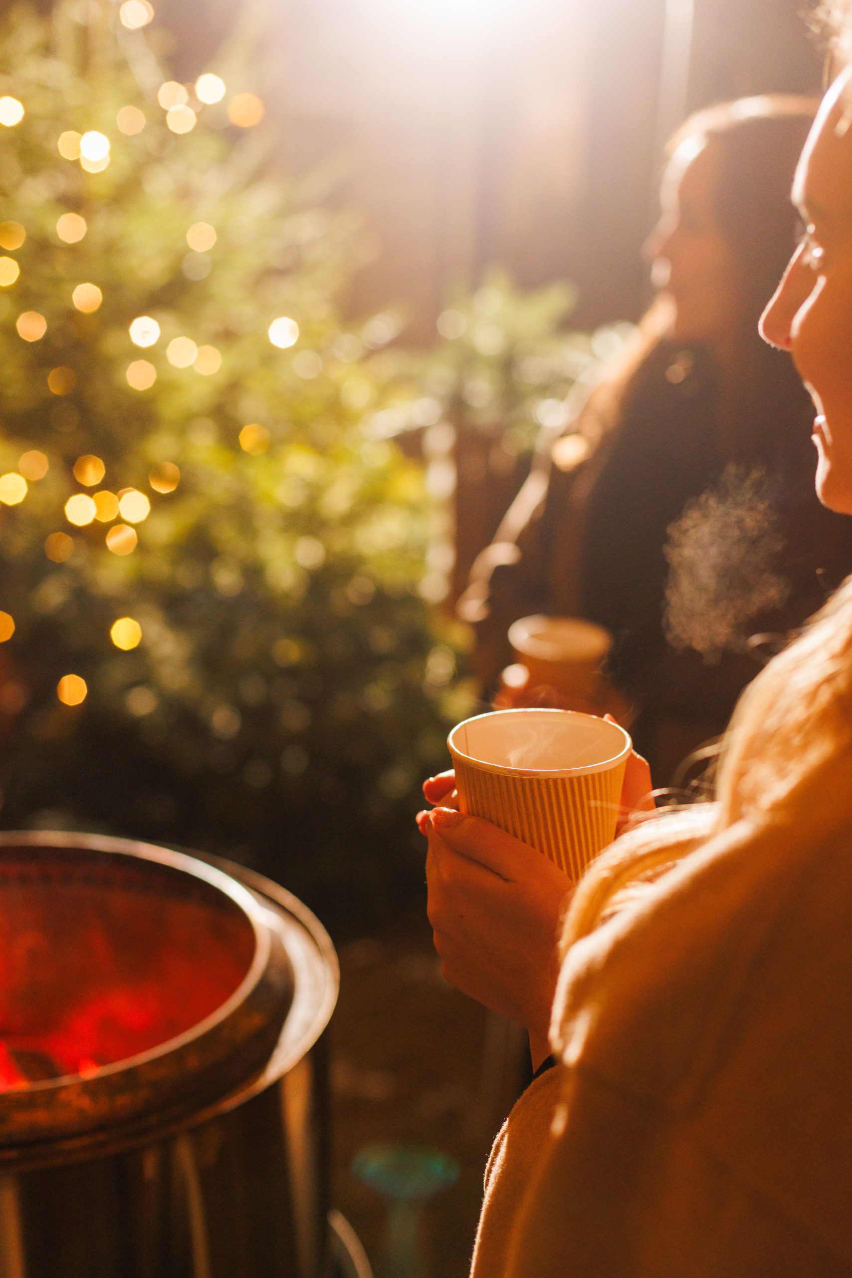 Zwei Personen halten heiße Getränke in gelben Tassen in der Nähe einer glühenden Feuerstelle im Freien. Der Atem der einen Person ist in der kalten Luft sichtbar. Im Hintergrund leuchten warmes Licht und unscharfe Weihnachtsbeleuchtung.