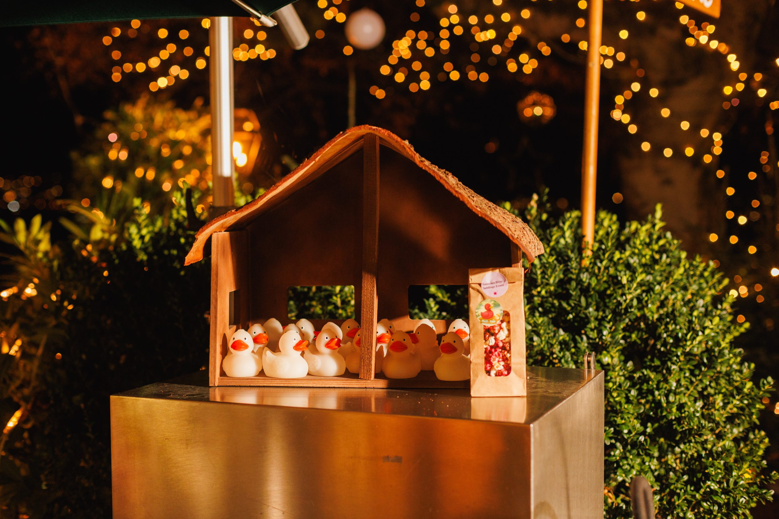 Ein kleiner hölzerner Krippenstall mit mehreren Gummienten steht im nächtlichen Freien, mit festlichen Lichterketten und Grünzeug im Hintergrund. Neben dem Stall steht eine Kiste mit Ornamenten.