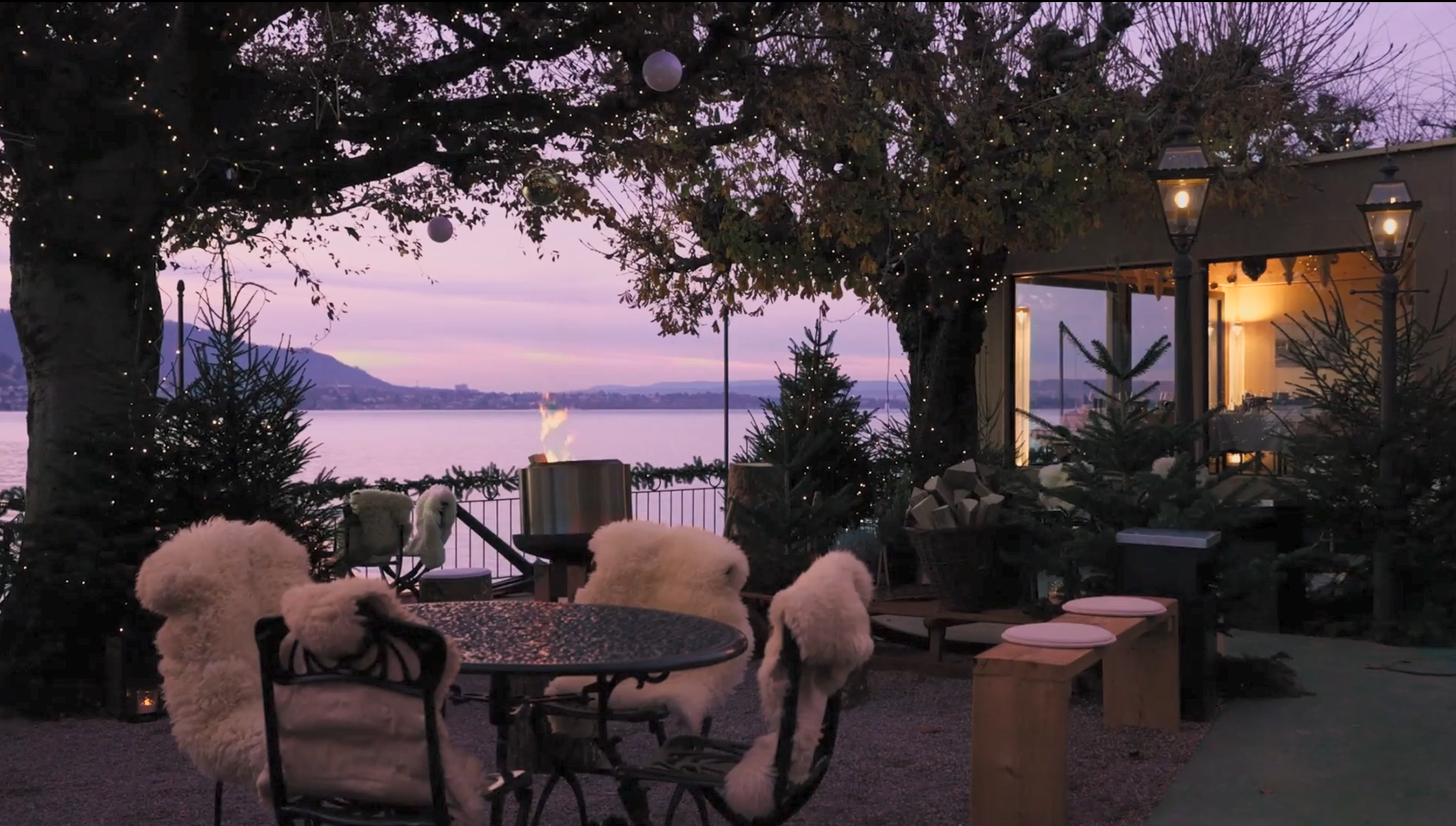 Eine Außenterrasse bei Sonnenuntergang mit Blick auf einen See und die Berge im Hintergrund. Gemütliche Stühle mit Fellüberwürfen umgeben die Tische, während Lichterketten von den Bäumen hängen. Im Chalet am See verleihen eine Feuerstelle und immergrüne Bäume dieser einladenden Szene Wärme.