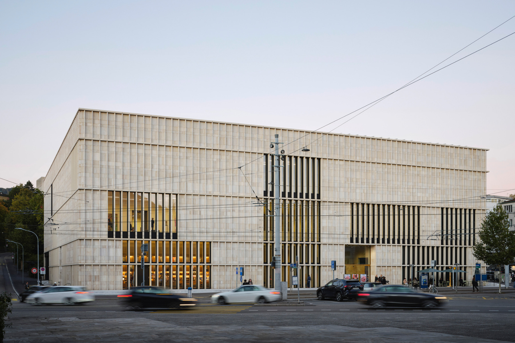 Un grand bâtiment moderne de forme rectangulaire avec une façade en panneaux de pierre claire, percée de hautes fenêtres verticales. Des voitures passent dans la rue devant et la scène est éclairée par une douce lumière naturelle - un endroit idéal pour une offre de loisirs.