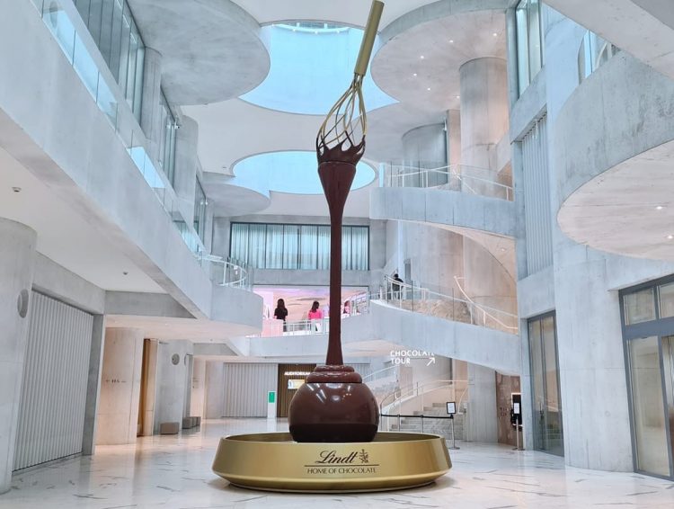 Une grande sculpture en chocolat représentant un fouet trempant dans du chocolat fondu se trouve au centre d'un atrium moderne et lumineux à la Lindt Home of Chocolate, une offre de loisirs entourée de balcons incurvés et de rampes en verre.