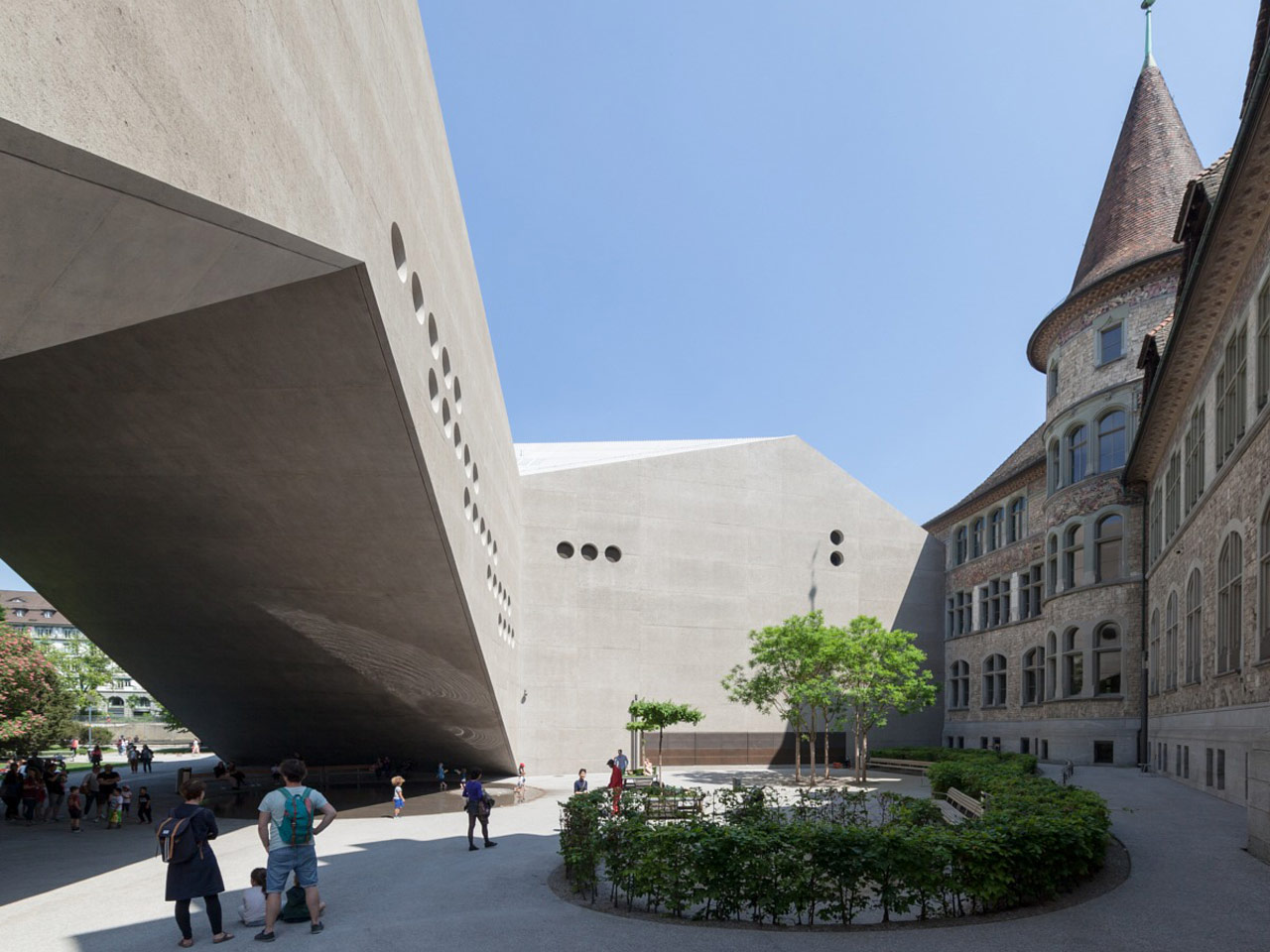 Des personnes marchent près d'un bâtiment moderne et anguleux en béton avec des fenêtres rondes, adjacent à un bâtiment historique en pierre avec une tour. Une petite place circulaire avec des arbres et des bancs offre un cadre idéal pour une offre de loisirs. Le ciel est clair et bleu.