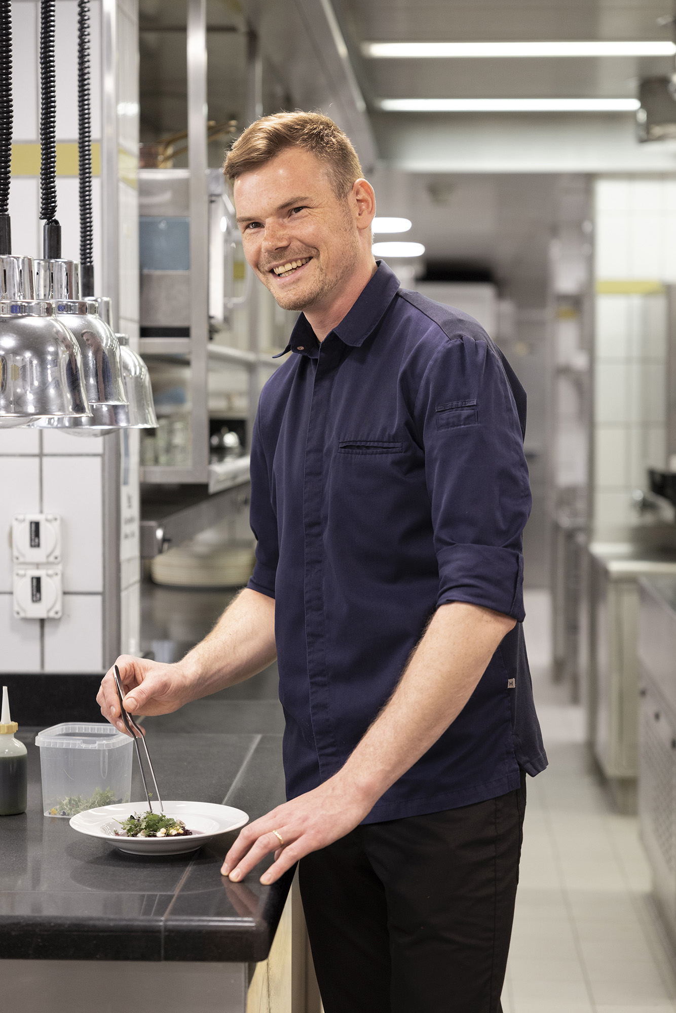 Un chef souriant, vêtu d'une chemise bleu foncé, utilise des pinces pour préparer des plats dans une cuisine commerciale moderne et lumineuse, debout à côté d'un comptoir où sont disposés divers ingrédients.