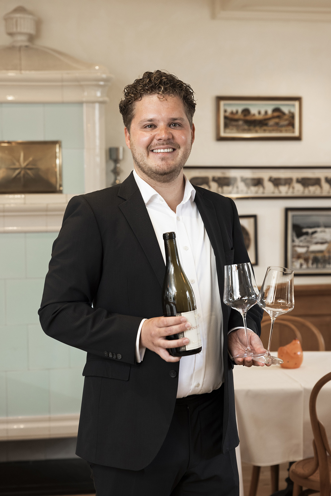 Un homme en costume noir est debout dans un restaurant, tenant une bouteille de vin et deux verres à vin, et souriant à l'appareil photo. Des photos encadrées et un poêle en faïence blanche sont visibles à l'arrière-plan.