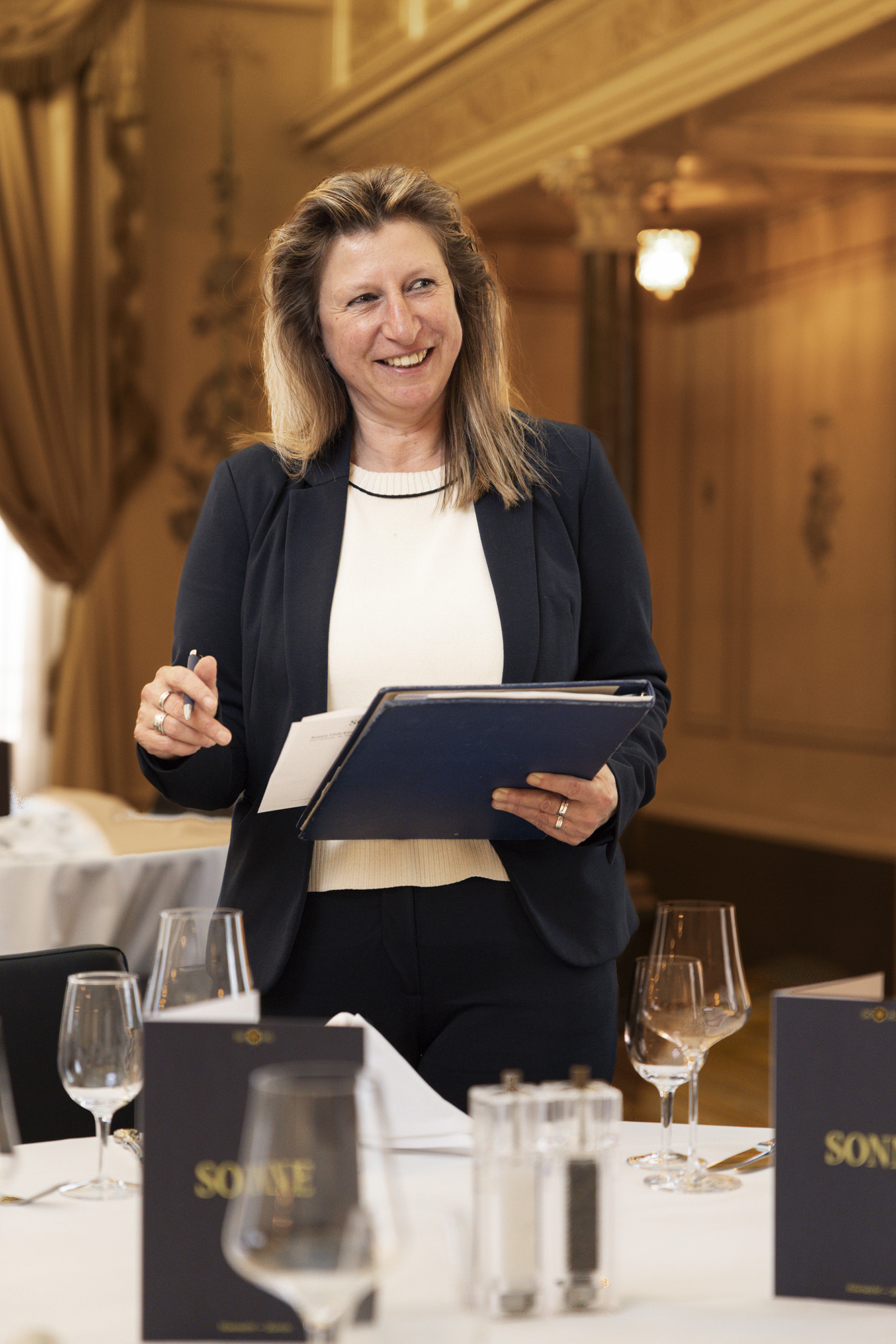 Une femme en tailleur sombre sourit dans un restaurant élégant, tenant un dossier et un stylo. Elle semble interagir avec les clients. Des verres à vin et des menus sont disposés sur une table devant elle.