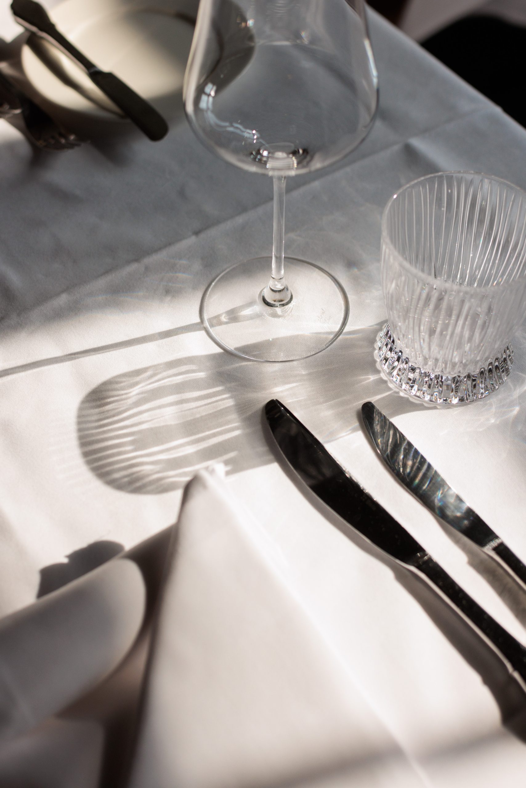 Nahaufnahme eines weißen Tischtuches mit einem Weinglas, einem Kristallbecher, Messer und Gabel im Restaurant am Zürichsee. Das Sonnenlicht wirft weiche Schatten auf den Tisch und faltet eine weiße Serviette im Vordergrund.