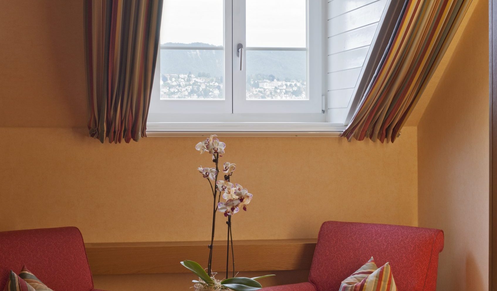 Zwei rote Sessel mit gestreiften Kissen stehen vor einem kleinen runden Tisch mit einer getopften Orchidee und zwei Äpfeln. Dieses einladende Hotelzimmer am See befindet sich unter einem weißen Bogenfenster mit gestreiften Vorhängen, das natürliches Licht hereinlässt.