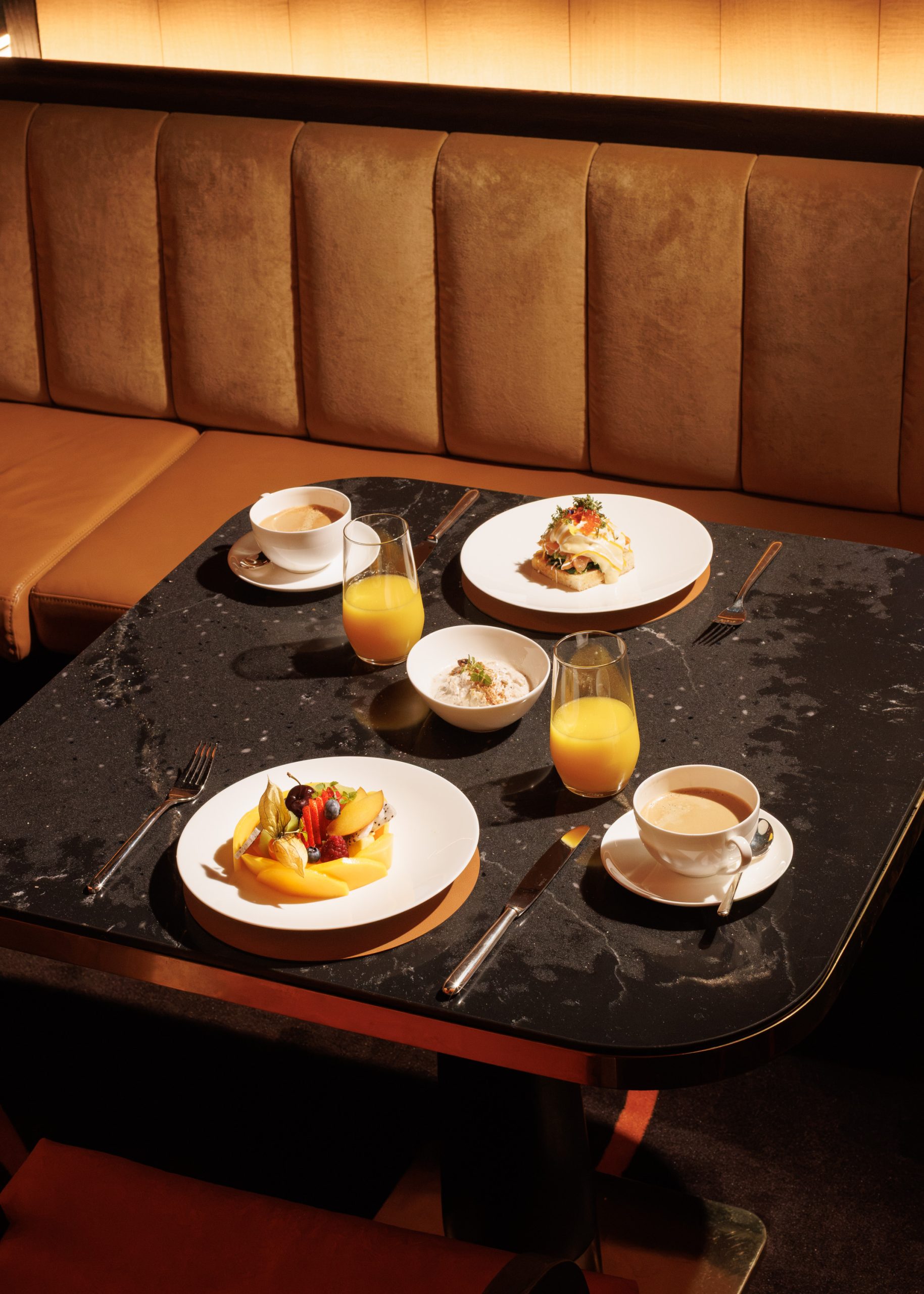 Auf einem schwarzen Tisch in der Nähe einer hellbraunen Sitzecke mit Polsterkissen erwartet die Gäste ein für zwei Personen gedeckter Restauranttisch mit Tellern voller Obst und Gebäck, zwei Gläsern Orangensaft, zwei Tassen Kaffee und einer Schüssel Joghurt – alles für ein köstliches Frühstück.