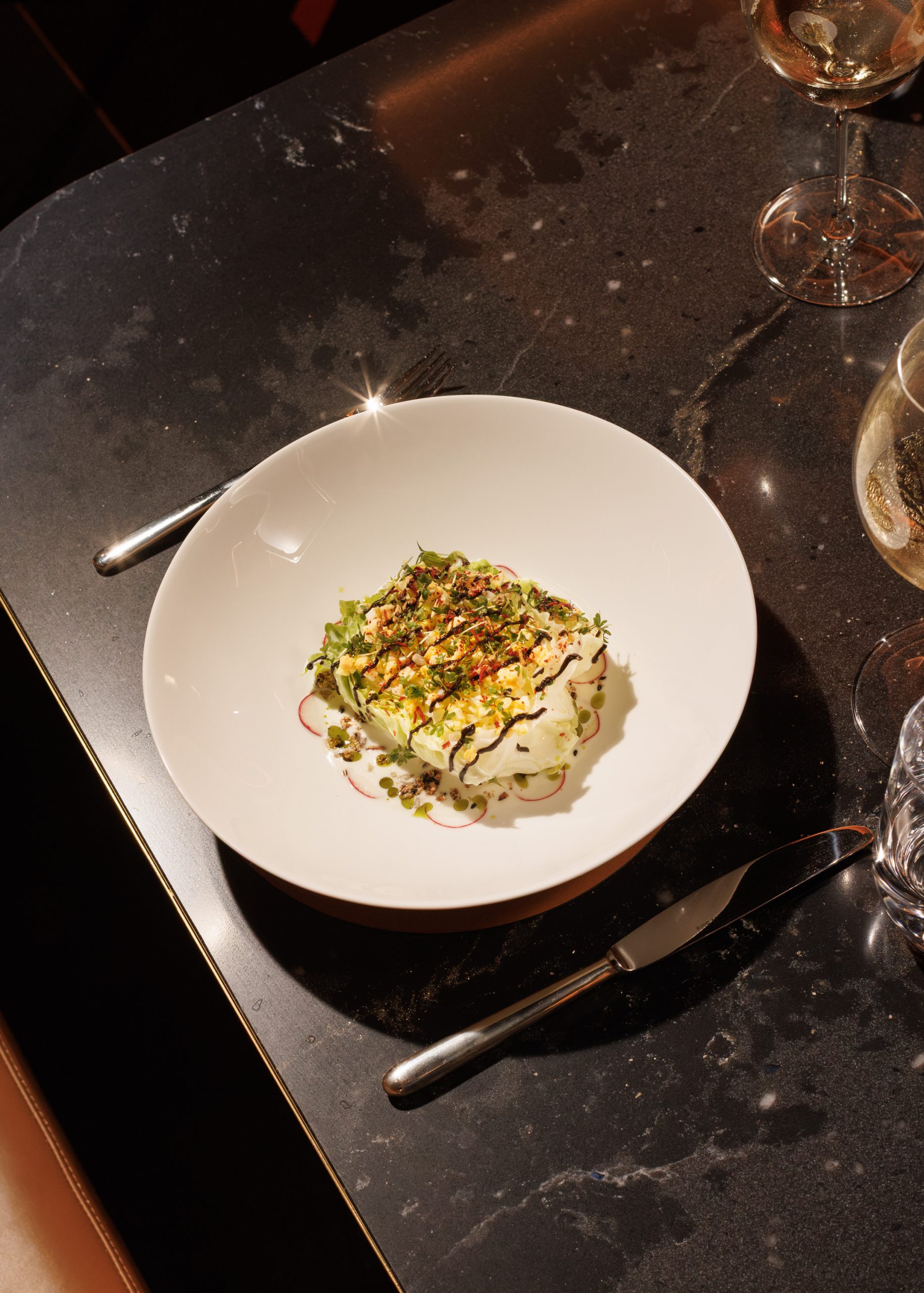Auf einem dunklen Tisch steht eine weiße Schüssel mit einem Gourmetgericht, das mit Kräutern und Soße garniert ist und zusammen mit einem Messer, einer Gabel und Gläsern mit Weißwein die Eleganz eines gehobenen Restaurants vermittelt.