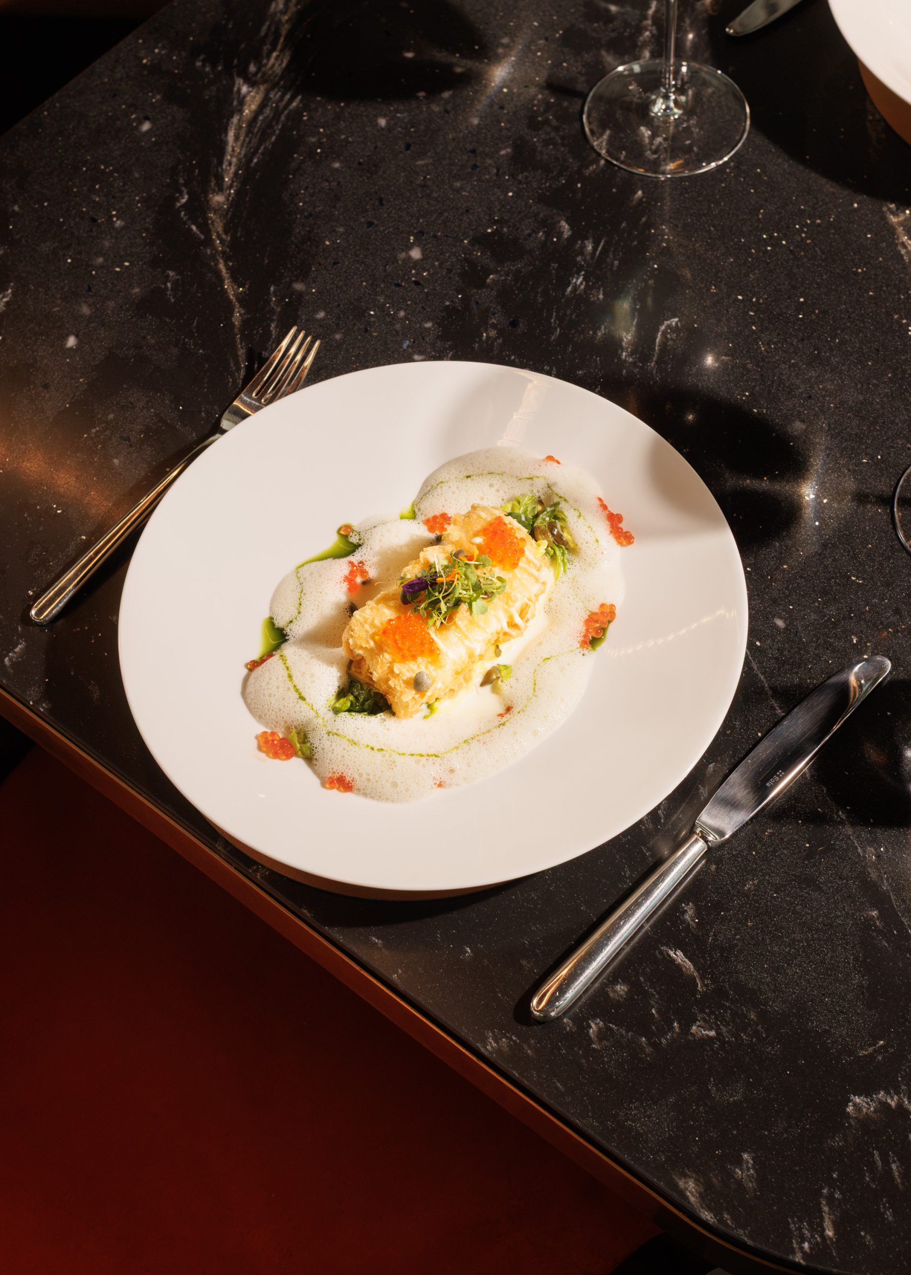 Ein ansprechend angerichtetes Gourmetgericht mit Schaum, Kräutern und Orangenrogen, serviert auf einem weißen Teller auf einem Tisch aus schwarzem Marmor in einem eleganten Restaurant. Daneben liegen Besteck und ein Weinglas.