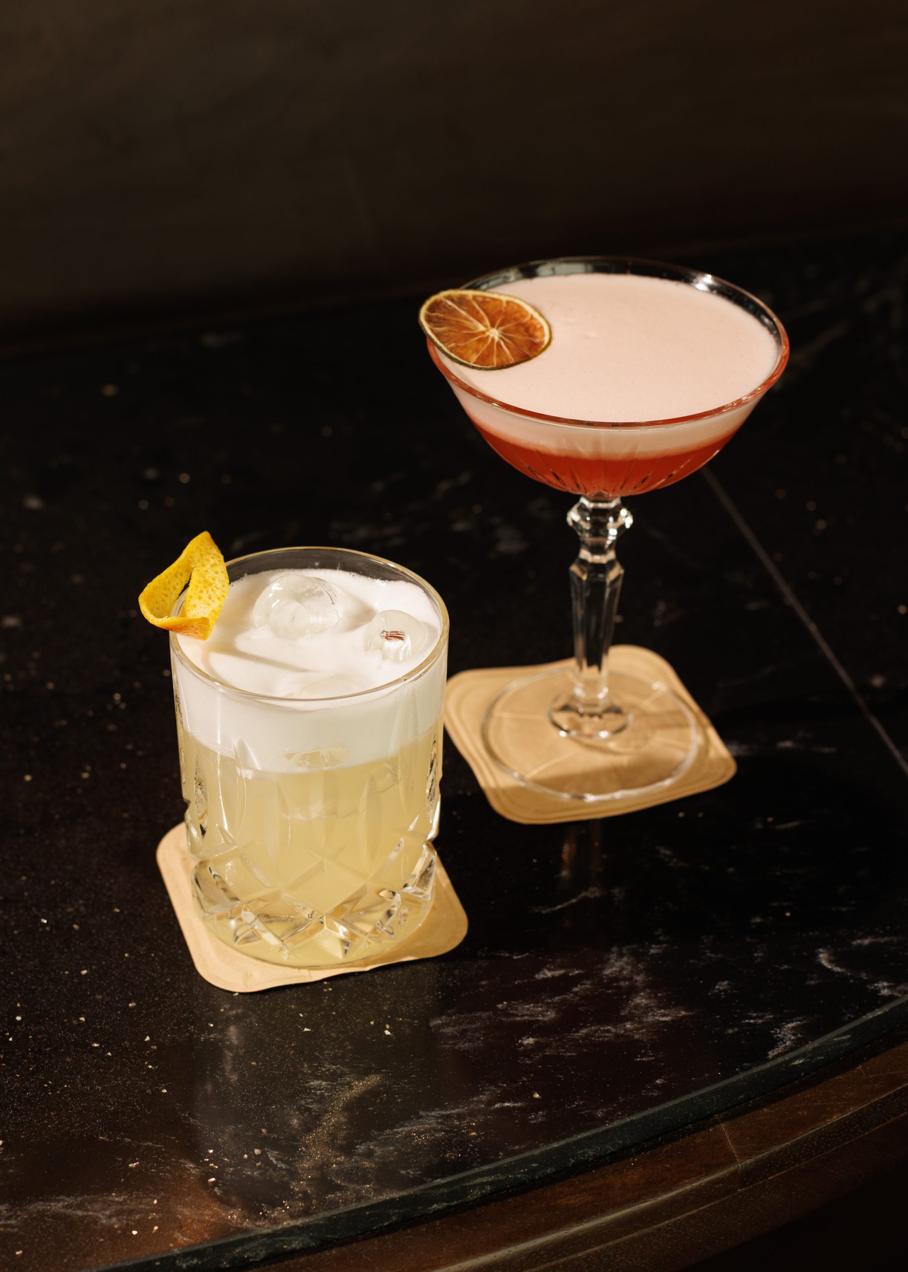 Zwei Cocktails auf quadratischen Untersetzern: einer in einem kurzen Glas mit Eis, Zitronenschale und Schaum; der andere in einem Coupé-Glas mit einem rosafarbenen Getränk und einer getrockneten Limettenscheibe als Garnitur. Beide stehen auf einem dunklen, glänzenden Restauranttisch.
