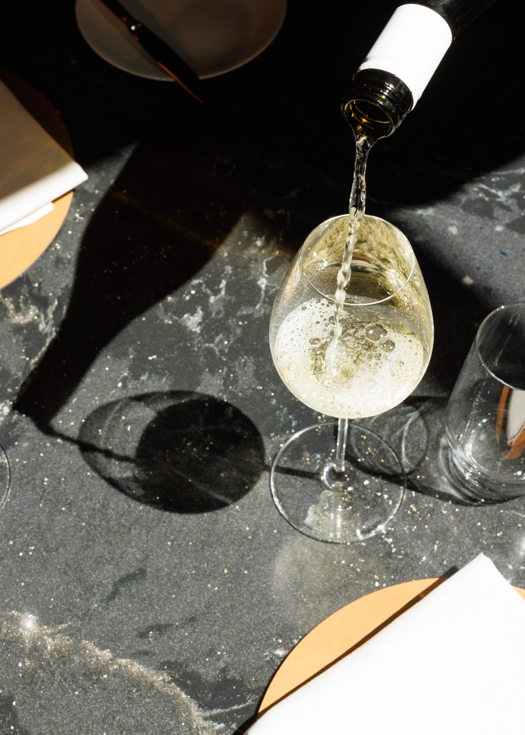 In einem Restaurant wird auf einem dunklen Marmortisch Weißwein aus einer Flasche in ein Glas gegossen. Man sieht Schatten und Bläschen, umgeben von Servietten, einem Teller und einem Trinkglas.