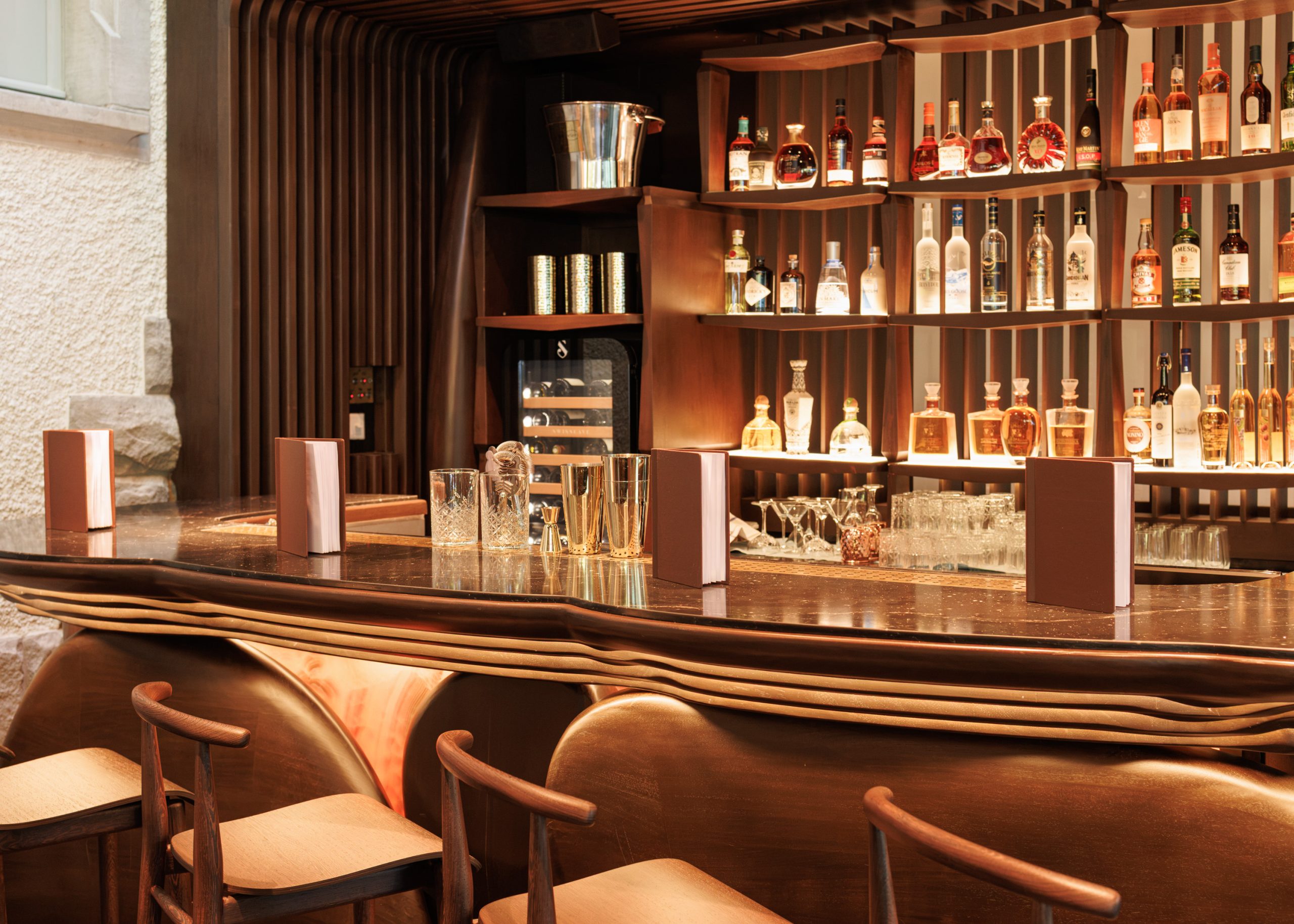 Eine elegante, moderne Bar in einem stilvollen Restaurant mit Holzhockern, einer polierten Theke, Spirituosenflaschen in beleuchteten Regalen, Cocktailgläsern und Speisekarten, die entlang der Bar ausliegen. Warme Beleuchtung sorgt für eine einladende Atmosphäre.