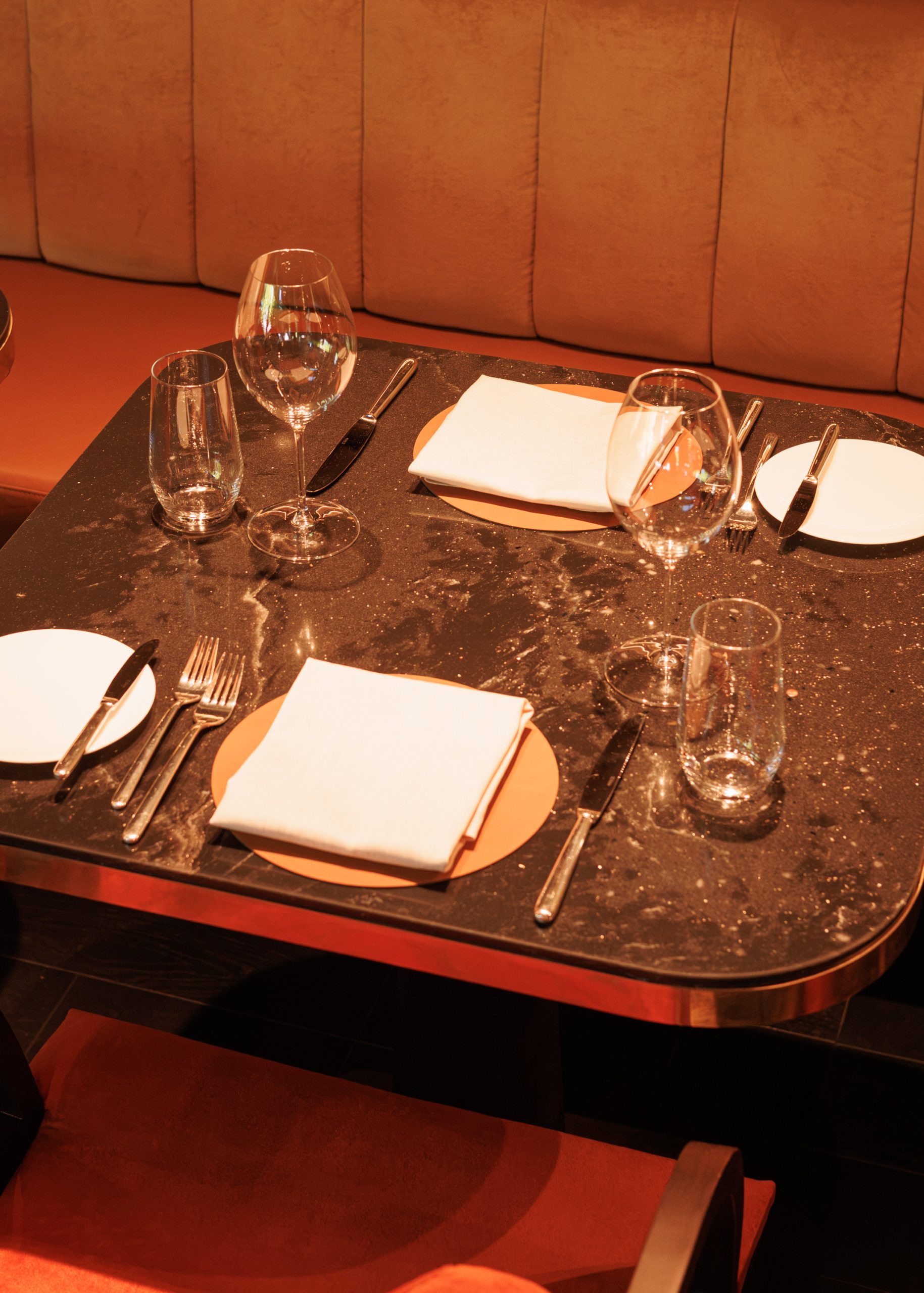 Ein gemütlich gedeckter Restauranttisch für zwei Personen mit Tellern, Servietten, Gabeln, Messern und Gläsern steht auf einer dunklen Marmorplatte, im Hintergrund ergänzen hellbraune gepolsterte Sitzgelegenheiten das Bild.