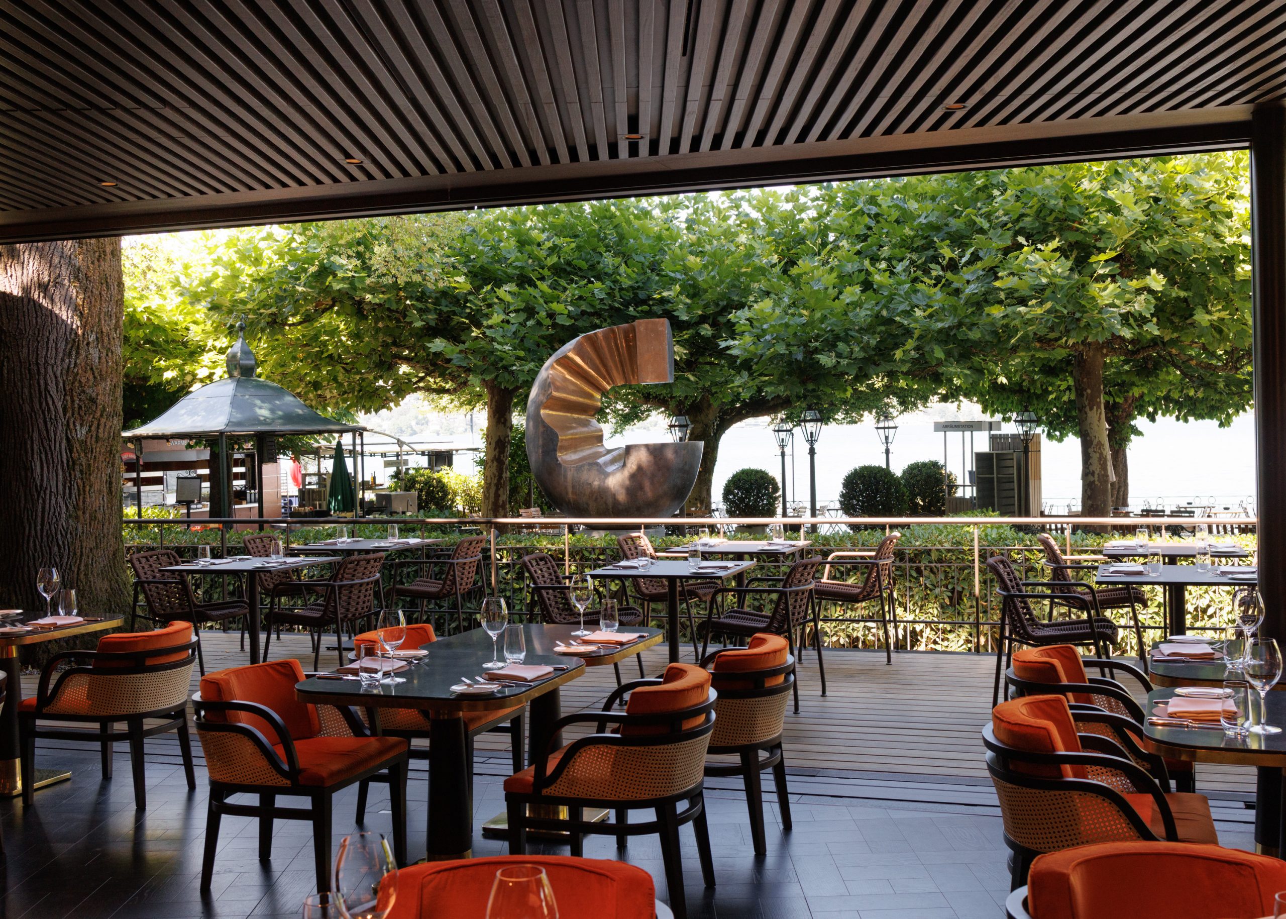 Die Sitzgelegenheiten im Außenbereich des Restaurants mit Holztischen und orangefarbenen gepolsterten Stühlen, von denen aus man auf einen Garten mit einer großen abstrakten Skulptur und üppig grünen Bäumen im Hintergrund blickt, schaffen eine lebendige und einladende Restaurantatmosphäre.