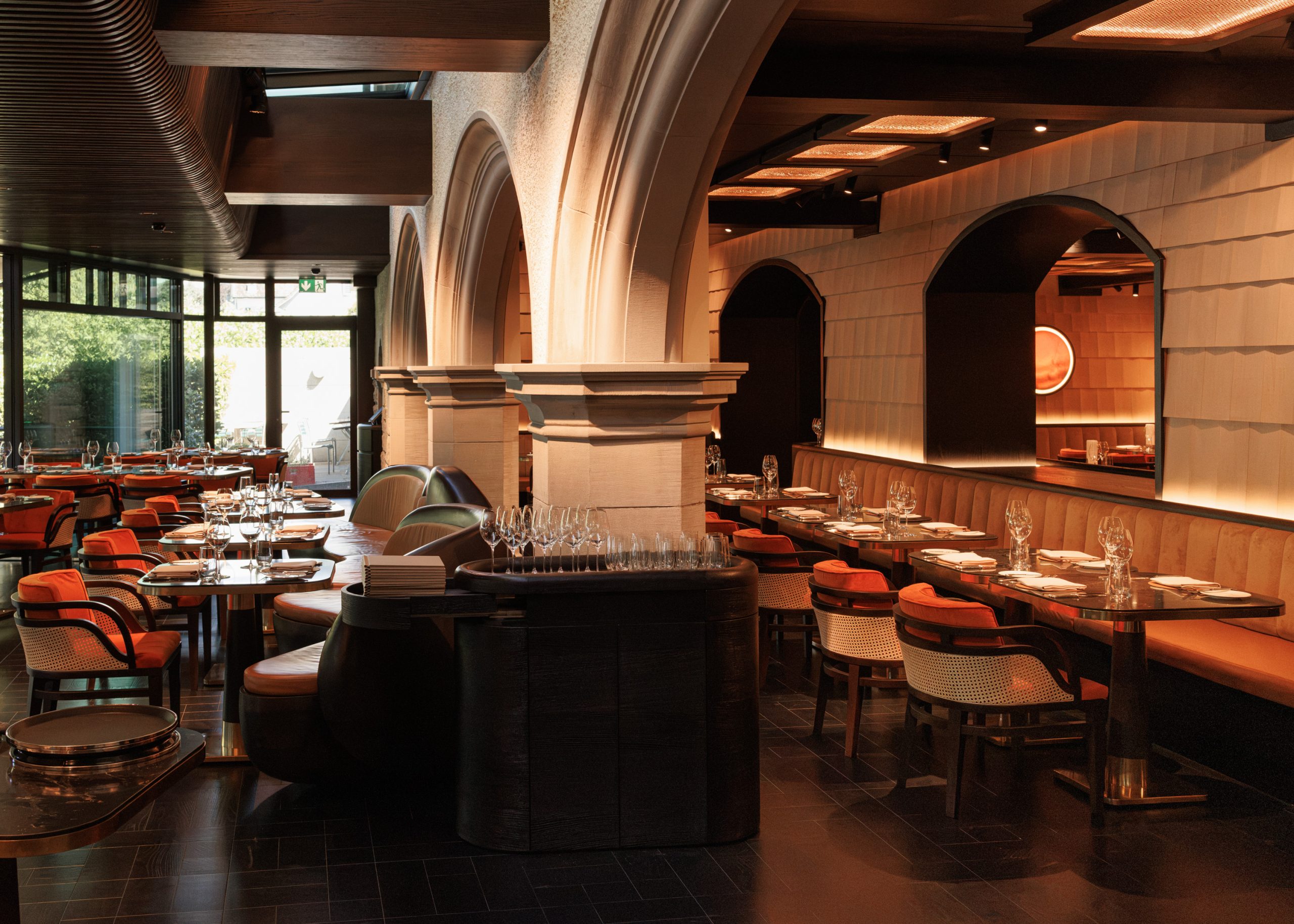 Elegantes Restaurantinterieur mit gewölbten Säulen, ordentlich gedeckten Tischen, modernen Stühlen mit orangefarbenen Kissen und warmer Stimmungsbeleuchtung. Große Fenster lassen Tageslicht herein, und der Restaurantbereich strahlt eine raffinierte, einladende Atmosphäre aus.