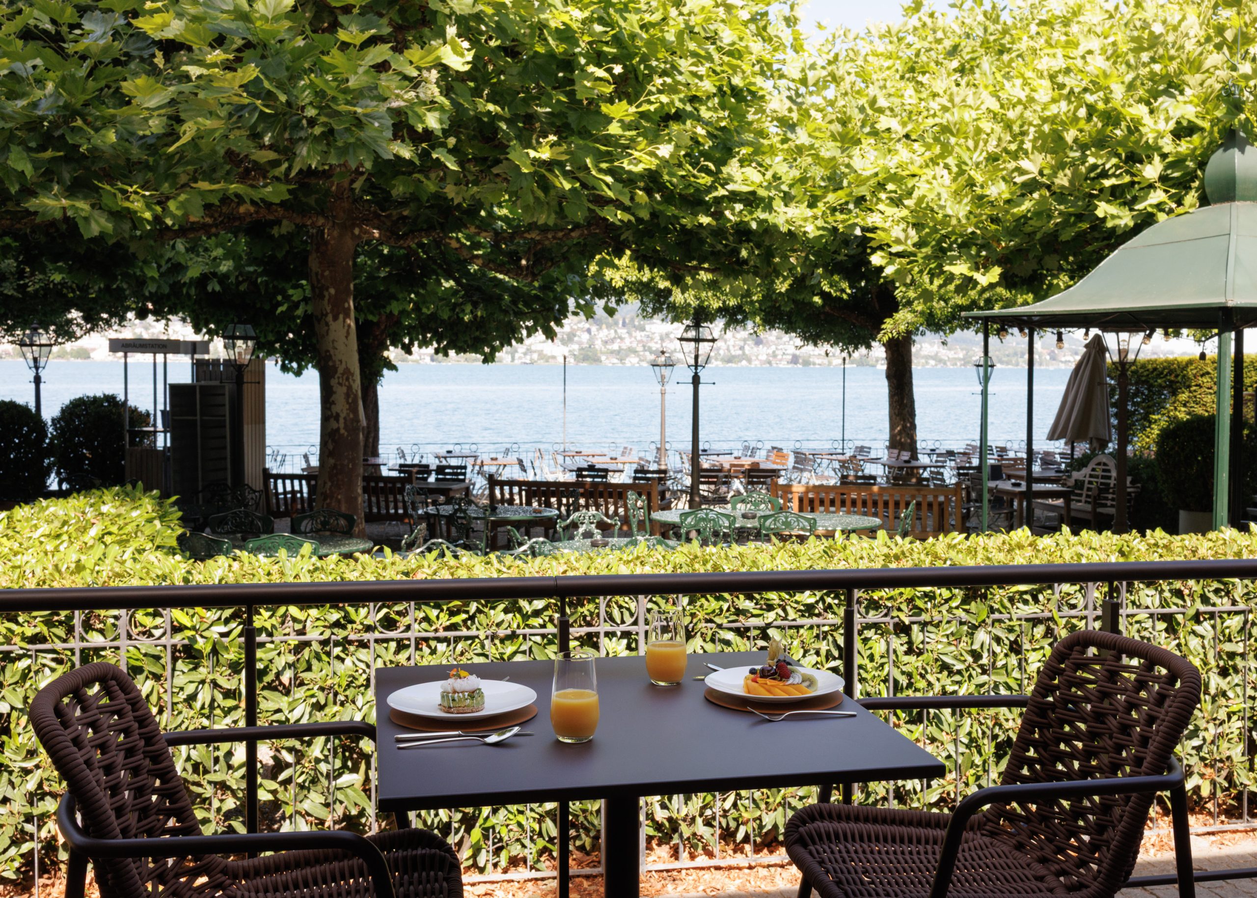 Ein für zwei Personen gedeckter Tisch mit Frühstück und Orangensaft bietet unter strahlend blauem Himmel einen Blick auf Reihen leerer Sitzplätze im Freien, üppige Bäume und einen See im Hintergrund.