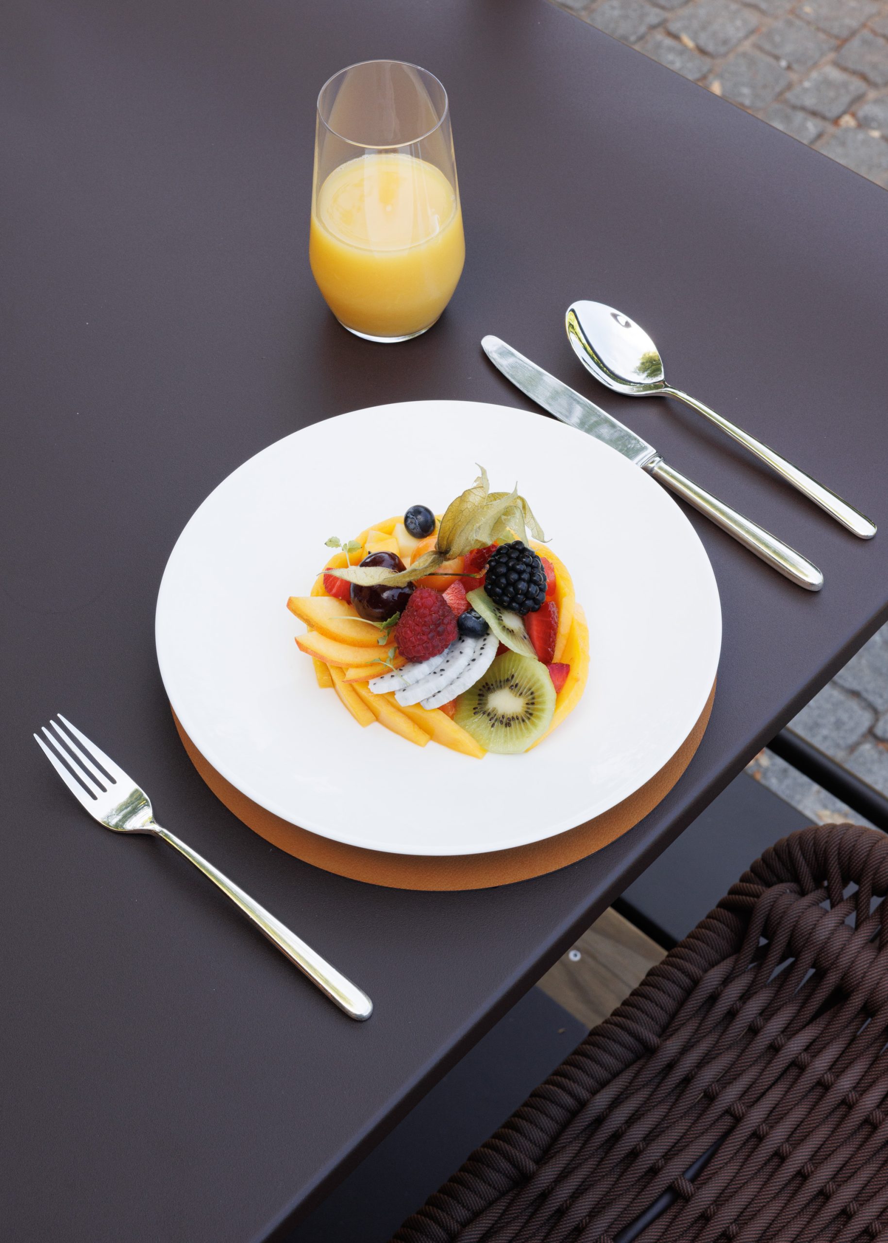 Auf einem braunen Tisch steht ein weißer Teller mit verschiedenen Obstscheiben und Beeren neben einer Gabel, einem Messer, einem Löffel und einem Glas Orangensaft – eine einladende Frühstücksszene. Ein geflochtener Stuhl und ein gepflasterter Boden sind teilweise zu sehen.
