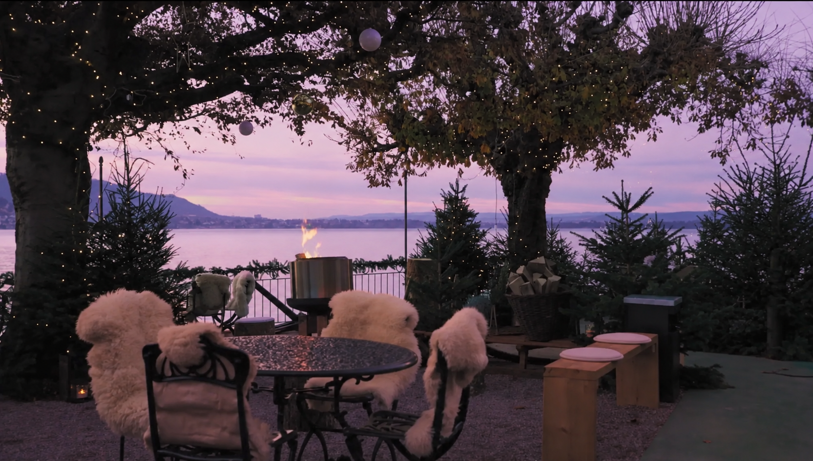 Terrassenbereich im Freien in der Abenddämmerung mit einer Feuerstelle, einem runden Tisch und mit Pelz bezogenen Stühlen, umgeben von kleinen Weihnachtsbäumen – für einen Winterzauber am Zürichsee mit Blick auf den See.
